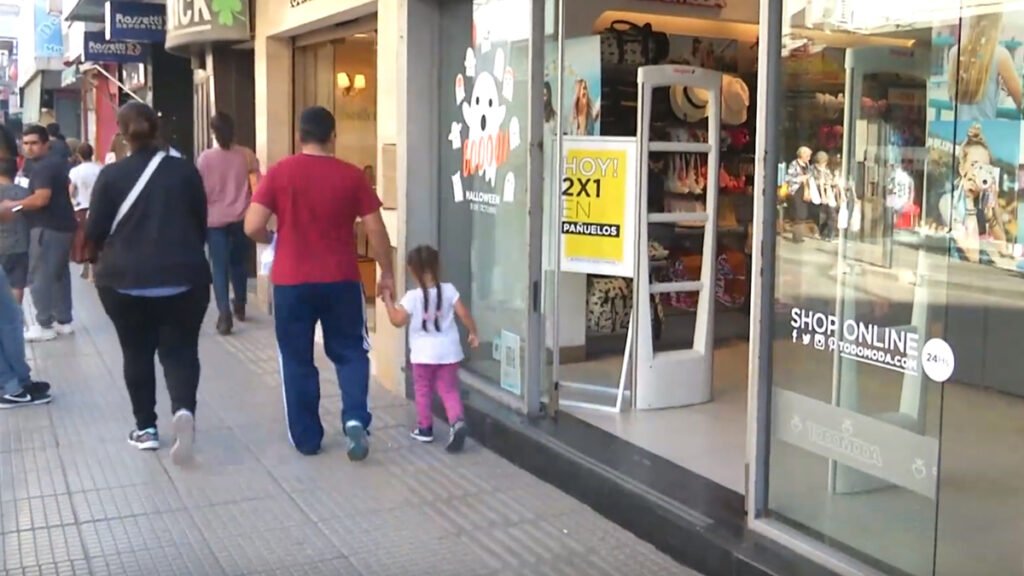 Esta imagen muestra a una familia caminando por la vereda frente a la vidriera de un local comercial con carteles de oferta y decoración de Halloween.La escena se ubica en una zona comercial urbana con circulación de peatones y escaparates iluminados.Se observa un letrero amarillo en la puerta del comercio que anuncia una promoción de 2x1 en pañuelos.